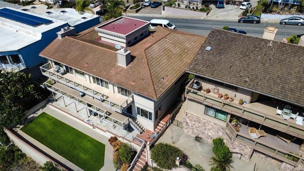 Aerial view of a multi-story residential property with a brown Spanish tile roof, multiple balconies, a flat rooftop structure, and a lawn, adjacent to a building with solar panels, with a street and cars in the background.