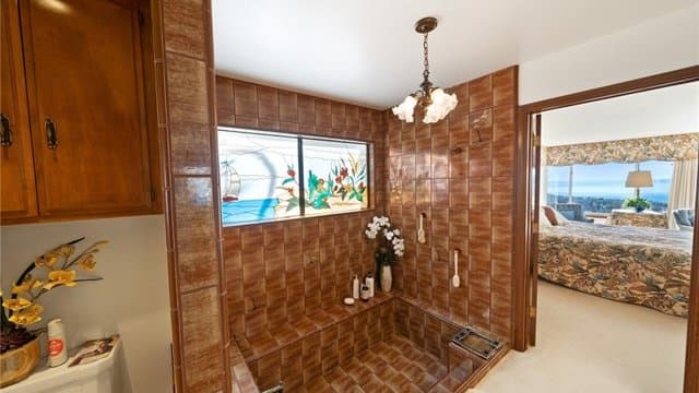A bathroom featuring extensive brown square tiles covering walls and a sunken bathtub, illuminated by a stained-glass window depicting a sailboat and coastal plants. An open doorway to the right reveals a bedroom with a panoramic ocean view.