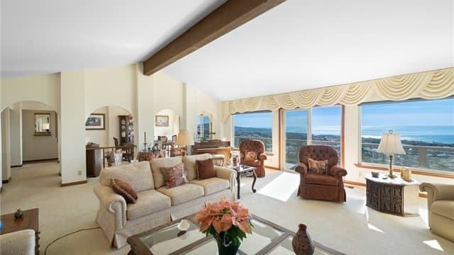 A spacious living room with a vaulted ceiling, light-colored sofa and armchairs, and large windows offering a panoramic ocean and coastline view. Arched doorways lead to adjacent rooms.