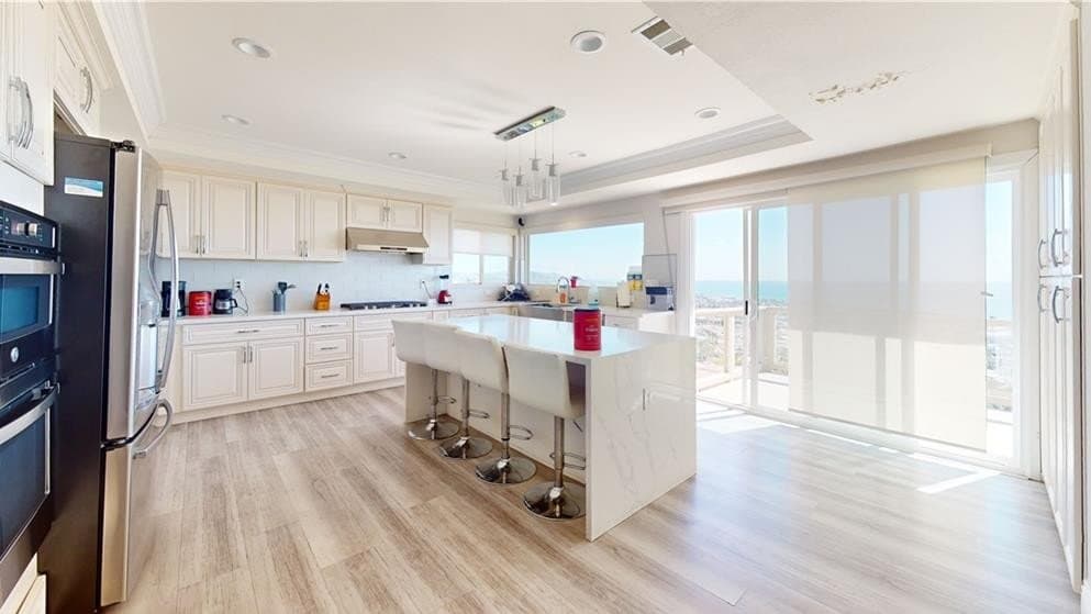 A bright modern kitchen with light cabinets, stainless steel appliances, a large white island with four white bar stools, and large windows looking out to a coastal view.