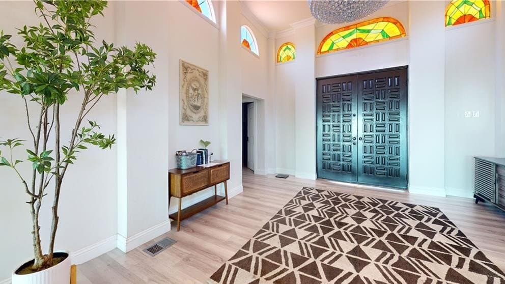 A grand entry hallway with high ceilings, several arched stained glass windows, a large dark double door with geometric patterns, a brown and white patterned rug, a tall potted tree, and a wooden console table.