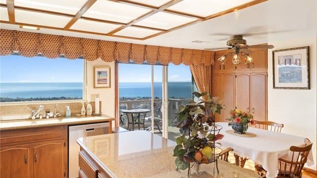 A kitchen featuring a central island with a quartz countertop, wooden cabinetry, a sink area, and a dining table, all set against a panoramic ocean view through large windows and a sliding door.