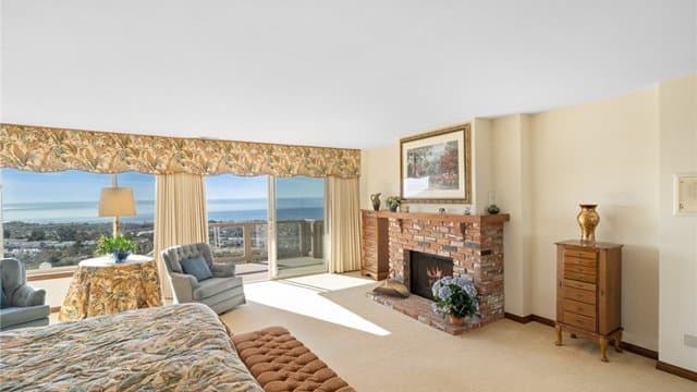 A spacious room featuring a bed with a patterned comforter, two armchairs, a small round table with a lamp, a brick fireplace, and large windows with a patterned valance that offer expansive ocean and city views.